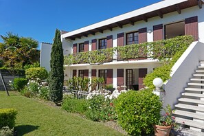 Um edifício de dois andares com uma varanda, rodeado por jardins bem cuidados e uma árvore alta.
