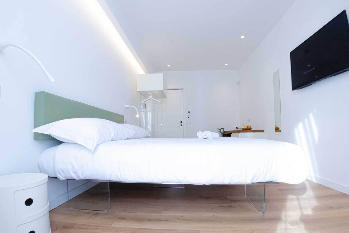 Ein modernes Hotelzimmer im minimalistischen Stil mit einem Bett mit grünem Kopfteil, einem weißen Nachttisch, einem an der Wand montierten Fernseher und Holzboden.