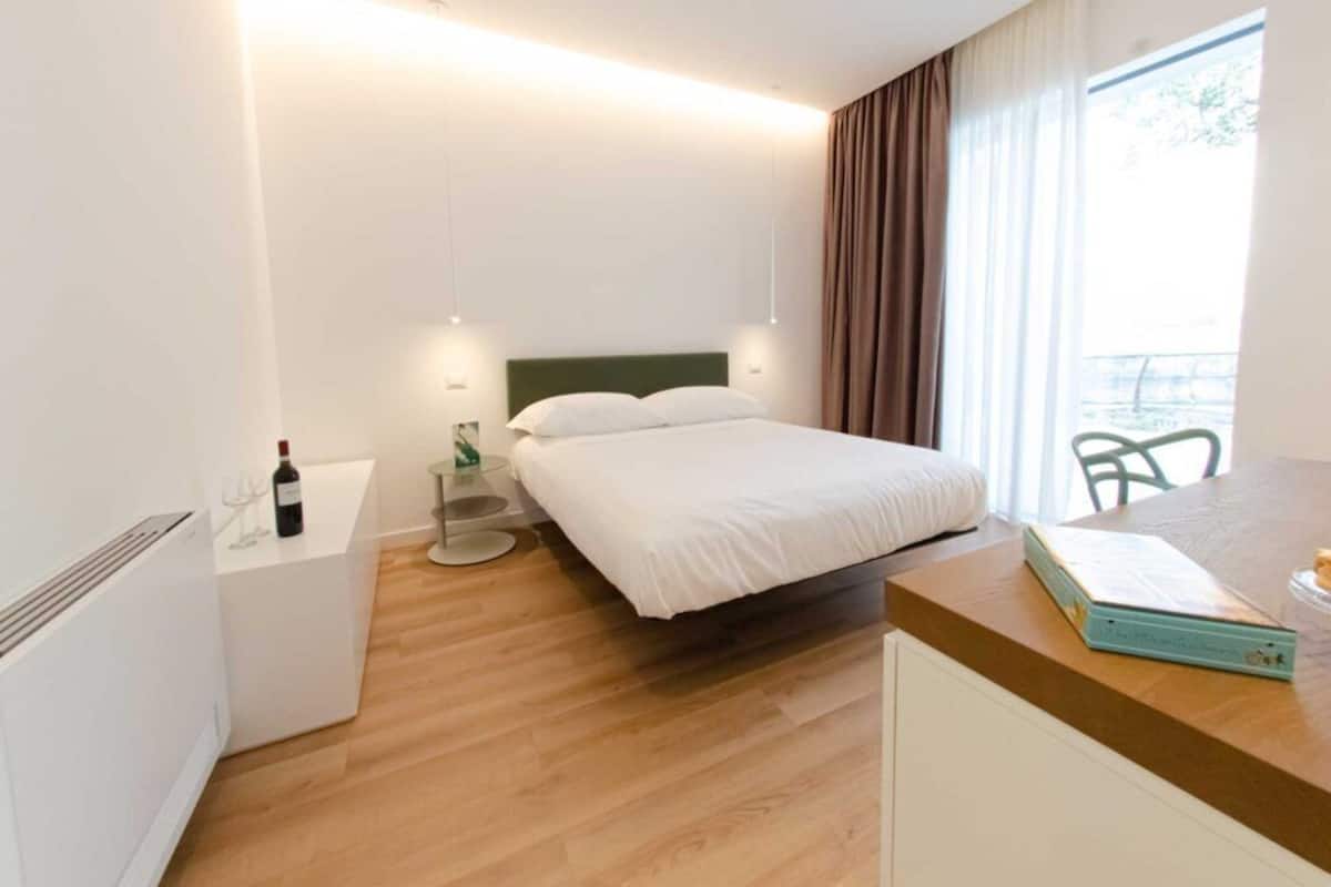 Ein Hotelzimmer mit einem Bett, einem Nachttisch mit Flasche und Gläsern, einem Sessel, einem kleinen Tisch und einem Fenster mit Vorhängen.