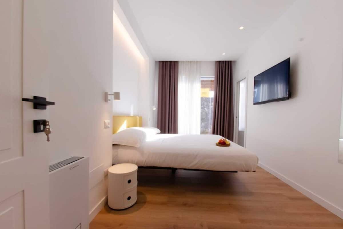 Ein modernes Hotelzimmer mit Bett, Wand-Fernseher, Nachttisch und einem kleinen weißen Papierkorb.