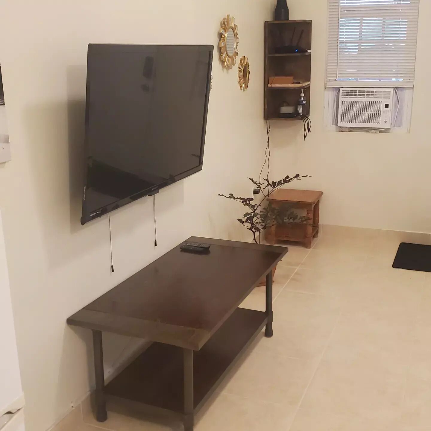 A living room with a flat-screen TV mounted on the wall, a dark wooden coffee table, and a small plant.