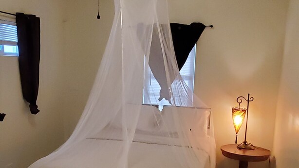 Ein Schlafzimmer mit einem Bett unter einem weißen Baldachin, einem kleinen Nachttisch mit einer Lampe, einem Fenster mit Jalousien und einem Deckenventilator.