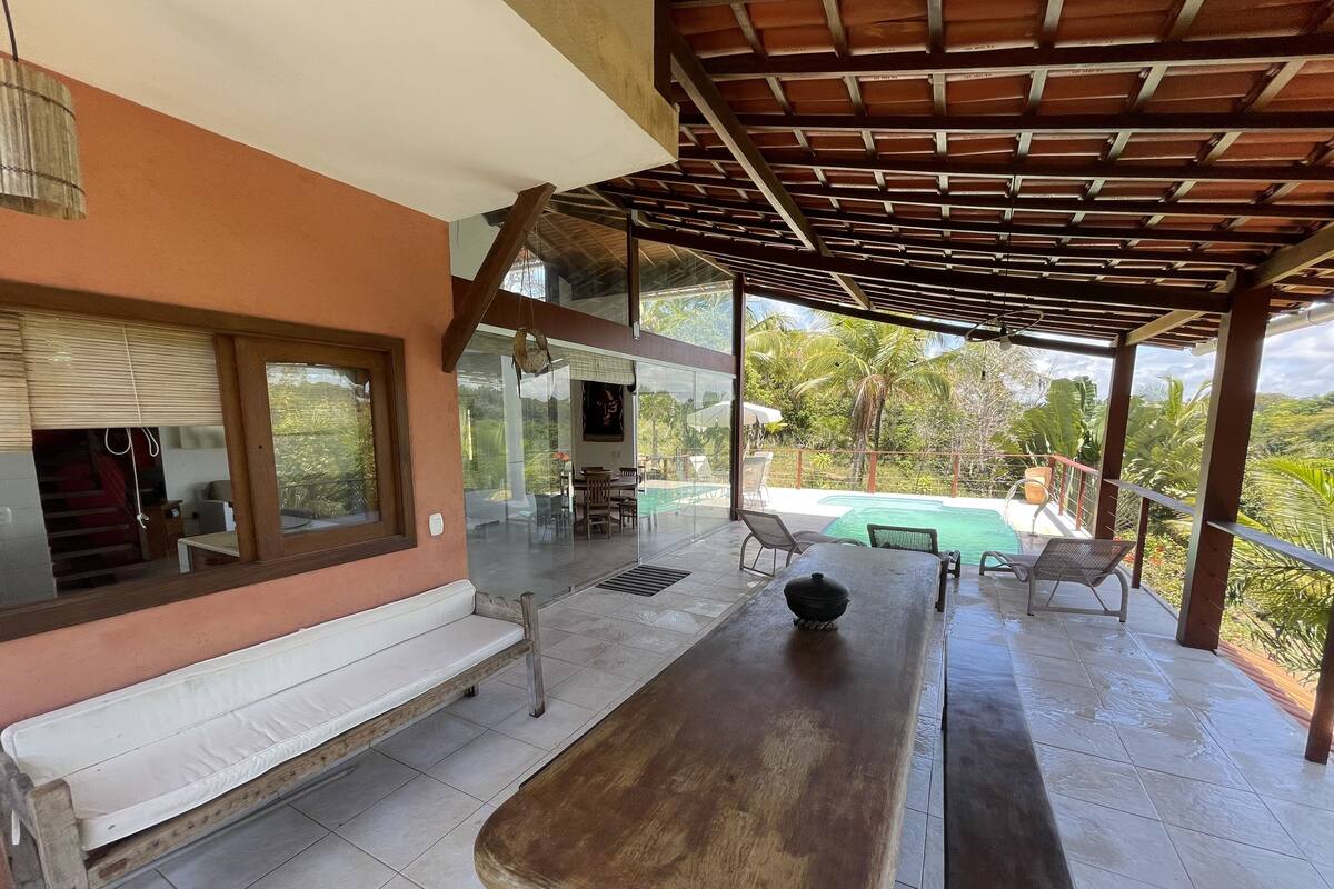 A covered outdoor seating area with a bench and table, overlooking a swimming pool and lush greenery.
