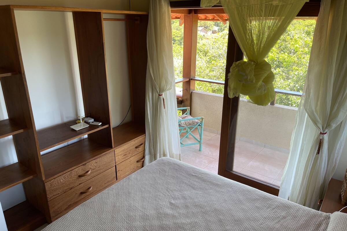 A bedroom with a wooden wardrobe, a bed, a small table with a chair, and a balcony with a view of greenery.