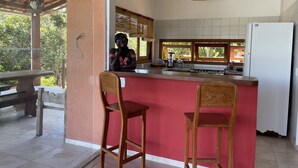 A kitchen with a red island, wooden chairs, a white refrigerator, and a window overlooking greenery. - Bia Trixx's Home (Porto Seguro)