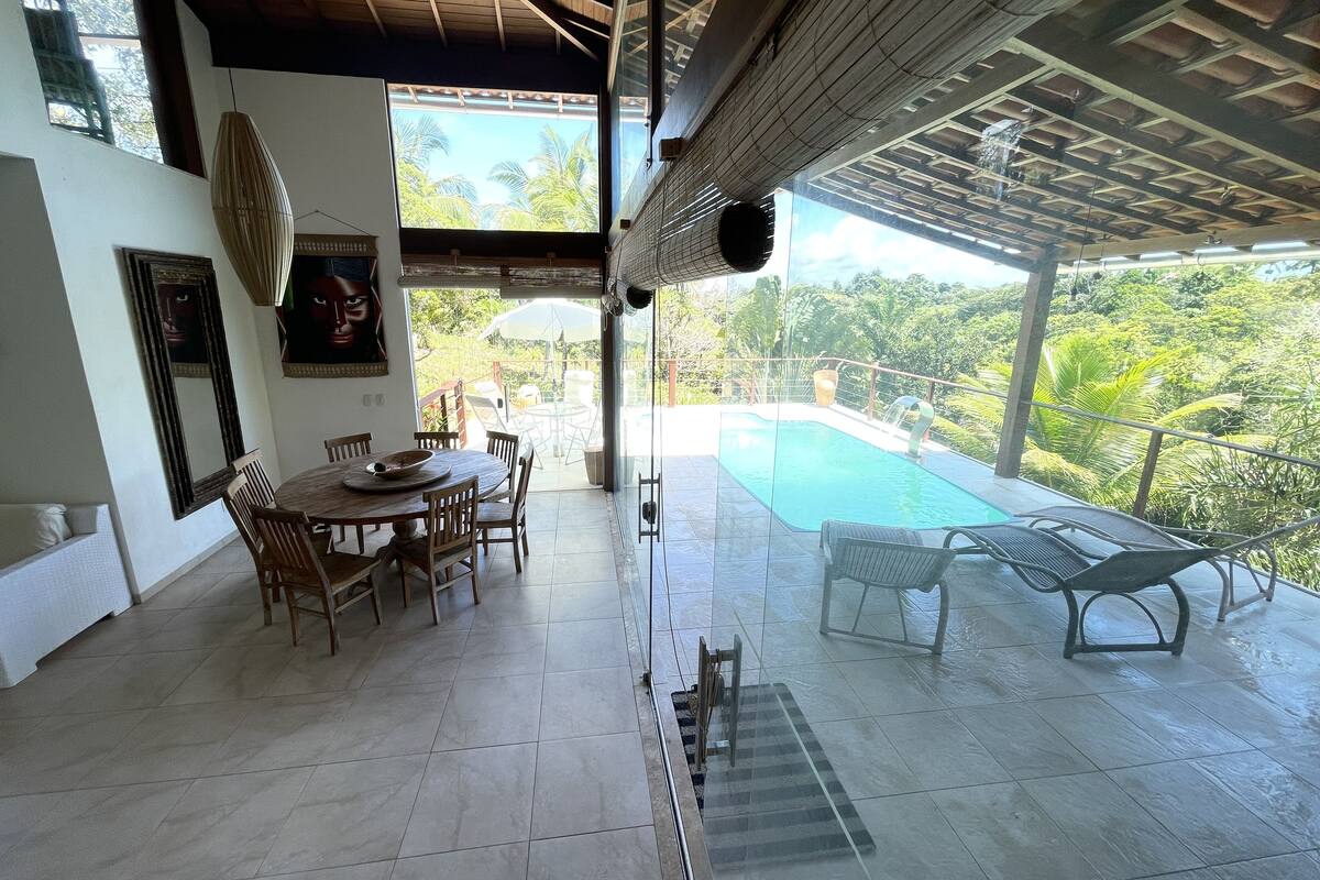 A dining area with a table and chairs, a large window, and a view of a swimming pool and lush greenery.