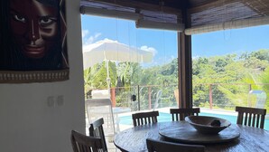 A dining area with a round wooden table and chairs, a large window overlooking a pool and lush greenery. - Bia Trixx's Home (Porto Seguro)