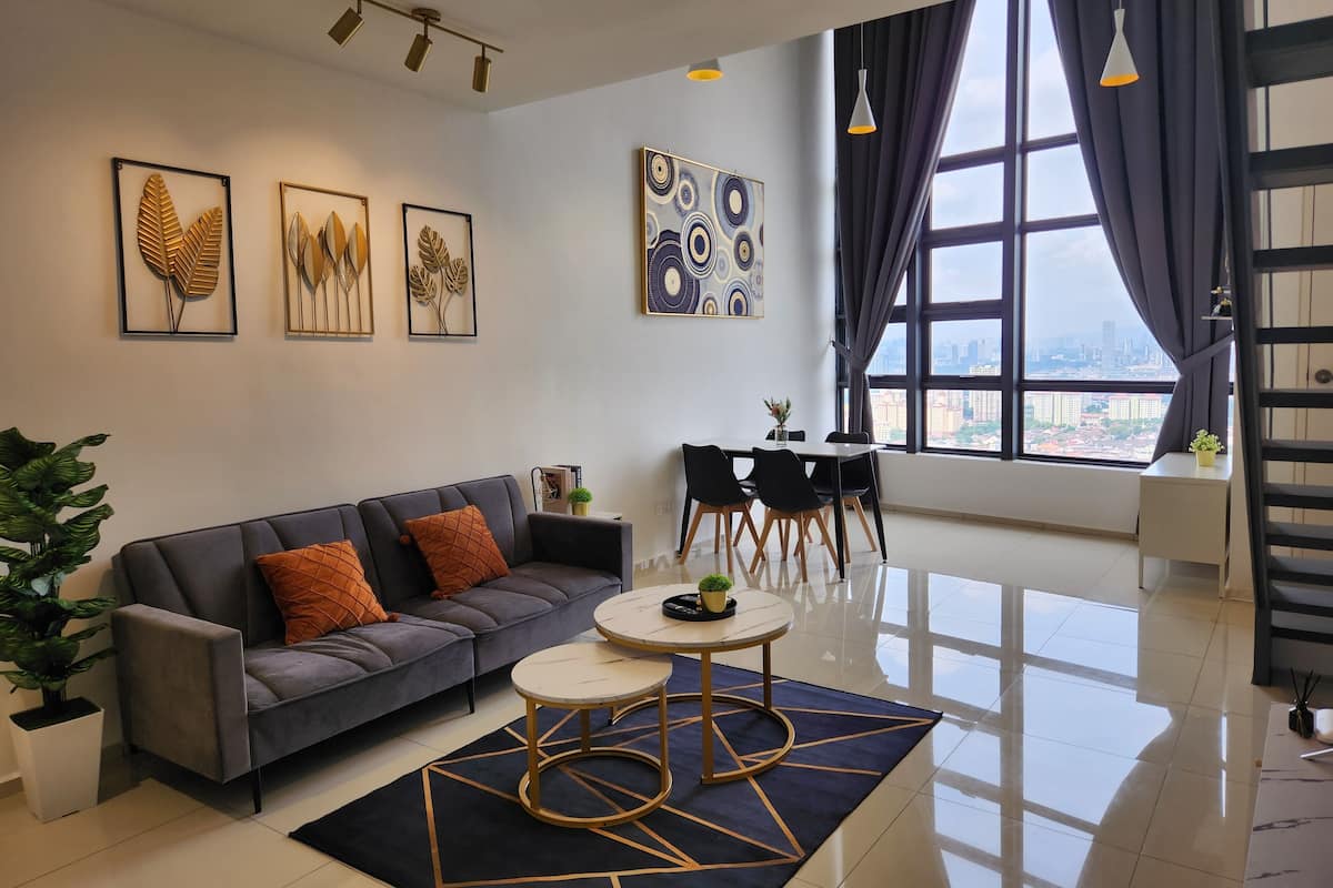 Una sala moderna con un sofá gris, una mesa de centro redonda y un comedor con una mesa blanca y sillas negras. Hay obras de arte enmarcadas en las paredes y un ventanal con cortinas.