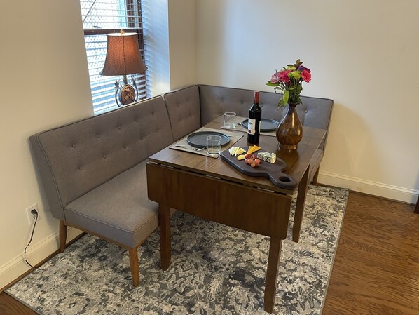 Un comedor con una mesa de madera, un banco acolchado gris, un jarrón con flores, una botella de vino y una tabla con queso y uvas.