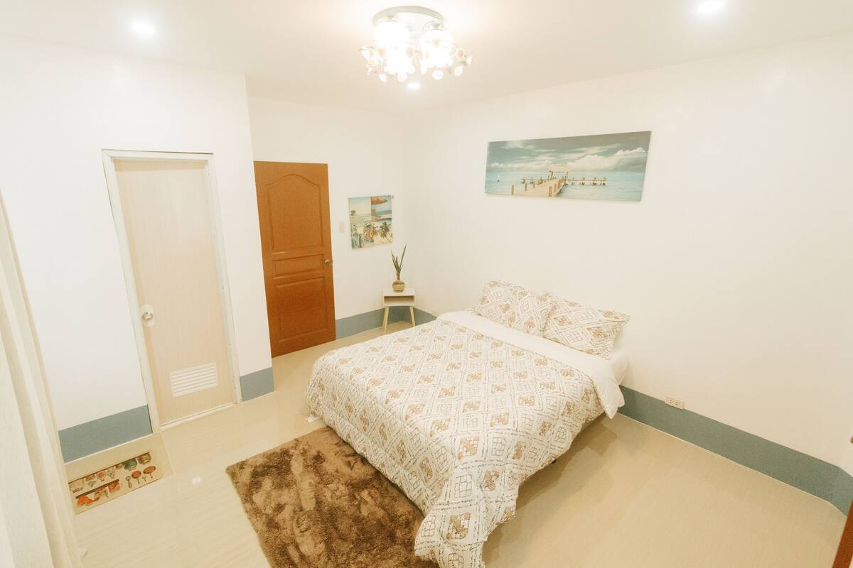 A bedroom with a bed, a door, a small table with a vase, and a painting on the wall.