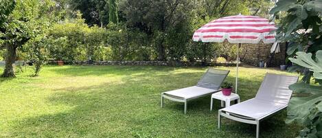 Un jardín trasero con césped, dos sillones blancos y un paraguas a rayas rojas y blancas.