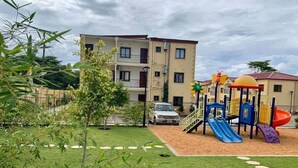 Ein Spielplatz mit Rutschen und Klettergerüsten, umgeben von einem eingezäunten Bereich mit Rasenfläche und einem Gebäude im Hintergrund.