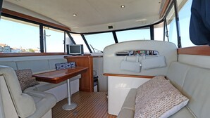 Interior de un barco con zona de asientos acolchada, una pequeña mesa y un televisor.