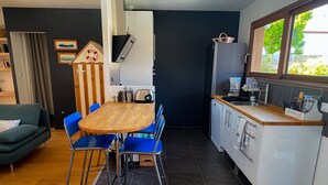 Eine kleine Küche mit einem Holztisch und blauen Stühlen, einem weißen Kühlschrank und einem Fenster mit Blick ins Grüne.