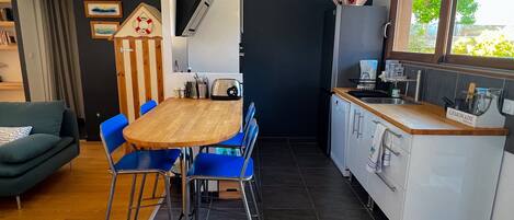 A compact kitchen with a wooden table and blue chairs, a white refrigerator, and a window with a view of greenery.
