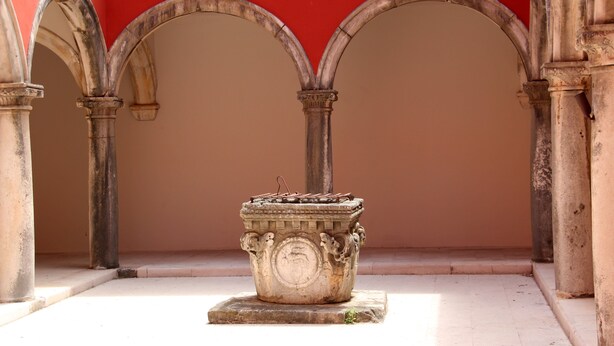 Un patio con una fuente de piedra, arcos y un muro rojo.