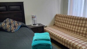 Una cama bien hecha con un edredón oscuro y una almohada decorativa, una mesita de noche con una lámpara, y un sofá a cuadros.
