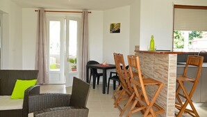 Un espace repas avec des meubles en osier, un coin bar avec des tabourets et une vue sur l’extérieur grâce à une porte vitrée.
