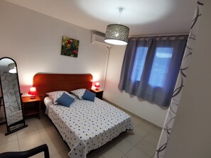 Quarto com cama, duas luminárias de cabeceira, espelho, ar-condicionado, janela com cortinas e uma obra de arte na parede.