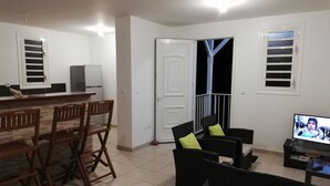 Un salon avec un téléviseur, un coin repas avec des chaises et une porte-fenêtre donnant sur un balcon.
