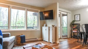 A living room with a sofa, a coffee table with books, a TV mounted on the wall, and a view of trees through large windows.