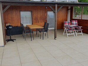 Une terrasse couverte avec une table et des chaises en bois, un barbecue et un coin salon agrémenté d’une couverture à carreaux.