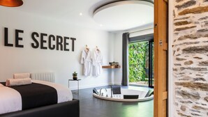 A modern bedroom with a bed, a wall-mounted sign reading "LE SECRET", and a bathroom with a bathtub and a view of greenery.