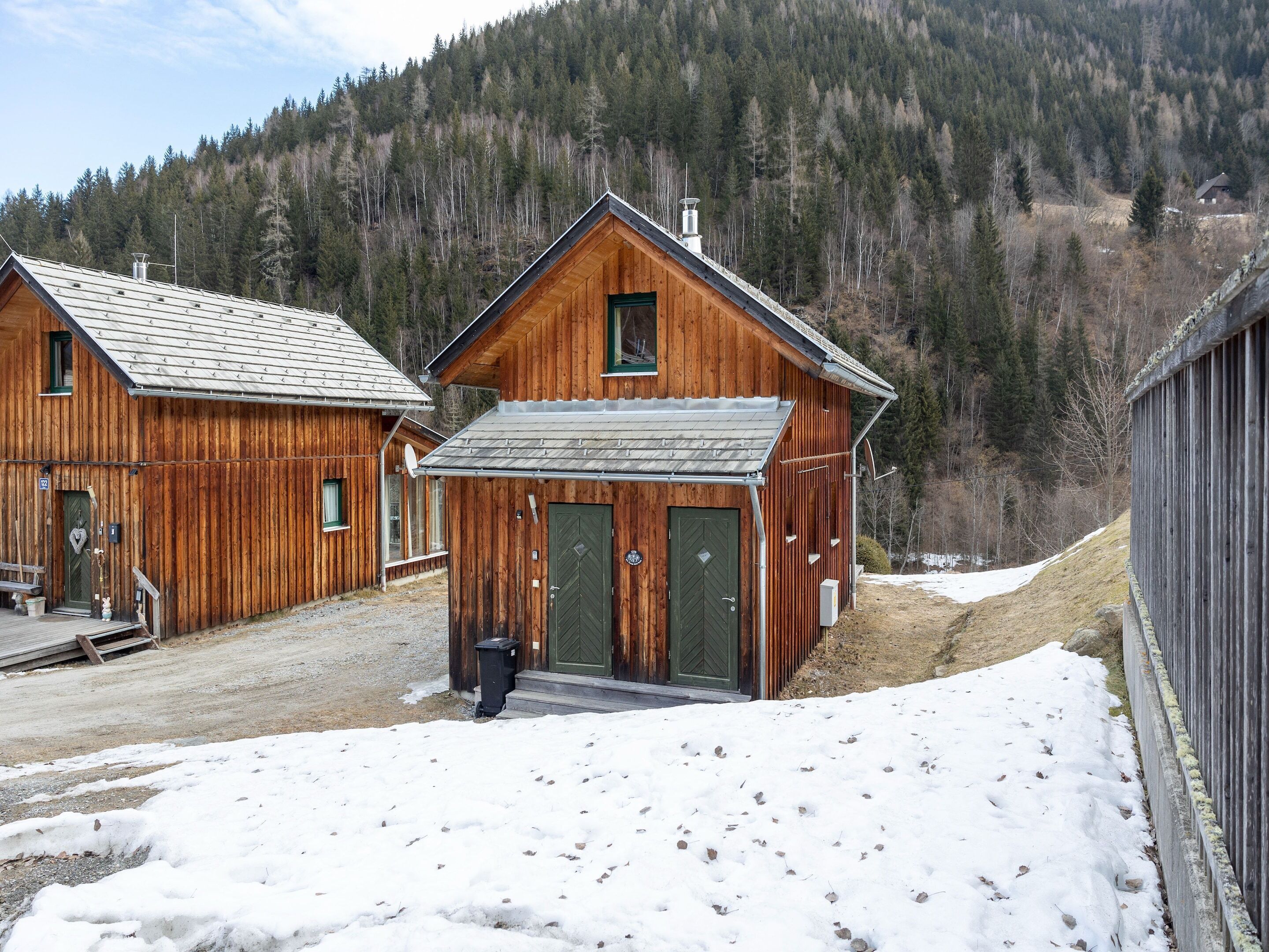 Eine Holzhütte mit grüner Tür und Metalldach, umgeben von verschneiter Landschaft und dichtem Wald.