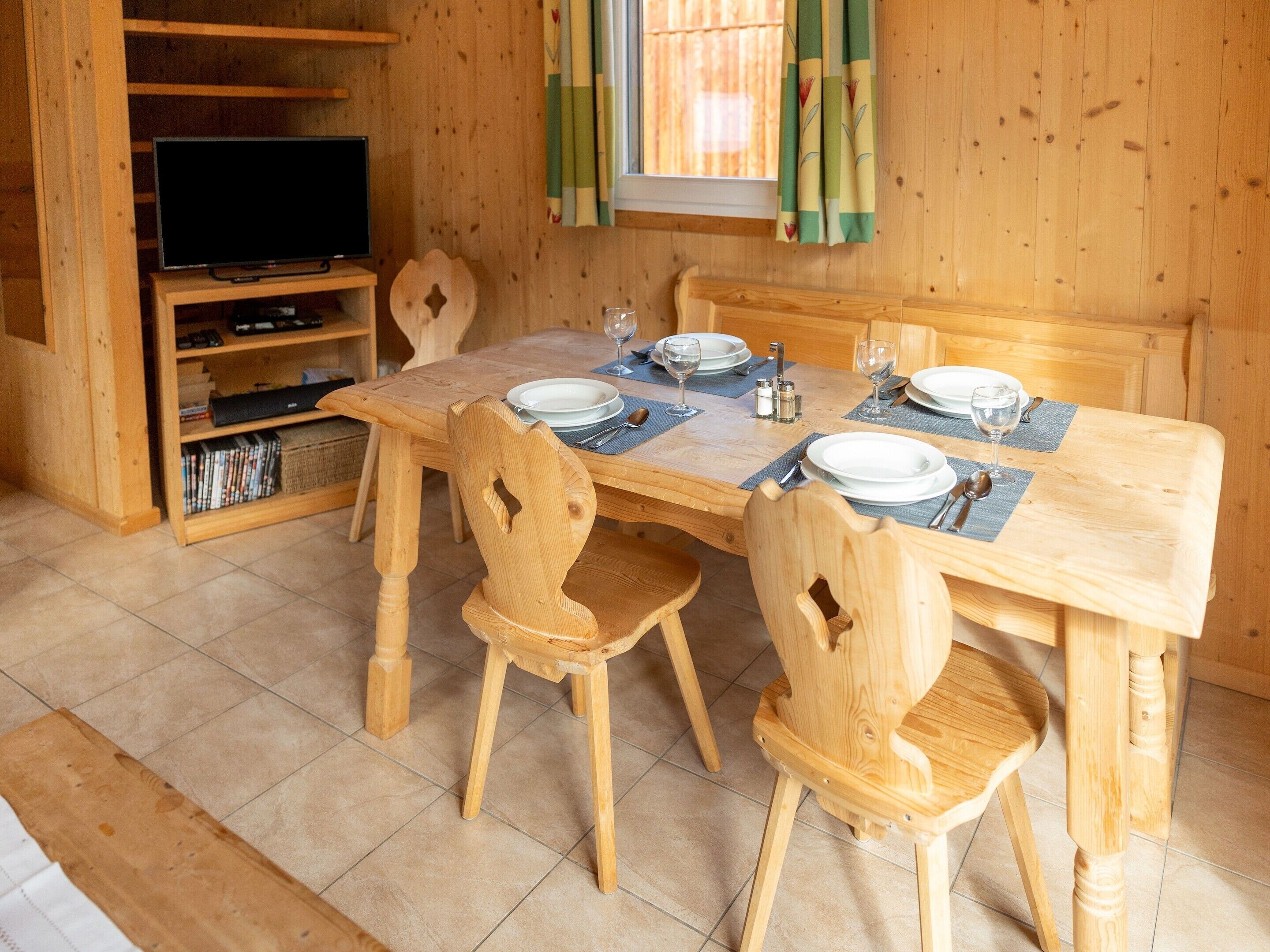 A wooden dining table set with plates, glasses, and cutlery, surrounded by wooden chairs.