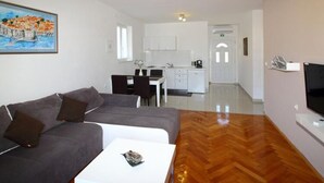 A modern living room with a sofa, a coffee table, and a TV. There is a kitchen area with cabinets and a dining table in the background. - Apartments Dubreta - Superior Two Bedroom Apartment With Balcony (Konavle)