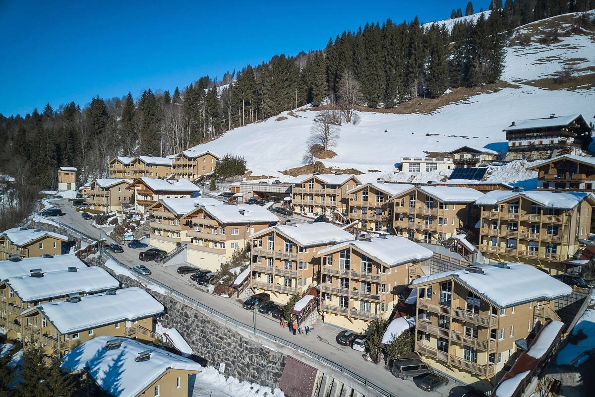 Un village de montagne enneigé, avec des bâtiments à plusieurs étages, un télésiège et une route.