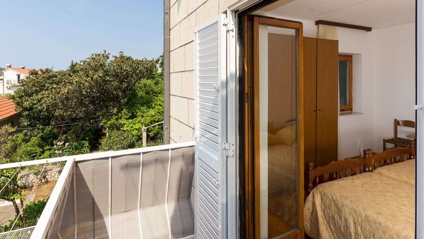 Un balcón con baranda metálica, un ventanal corredero que da a un dormitorio con cama, una silla de madera y un ventanuilla.