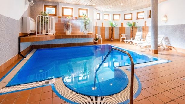 Piscina interna com teto de madeira, paredes azulejadas e um ambiente iluminado por sol, com espreguiçadeiras e plantas em vasos.