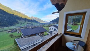 A balcony with a view of a valley and mountains, a small table with chairs, and a window with a view of a house. - Apartment in Neustift Near Ski Lift (Neustift Im Stubaital)