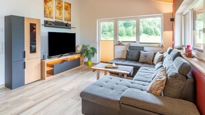 Een moderne woonkamer met een grote flatscreen-televisie, houten balken in het plafond, een zithoek en een eettafel.