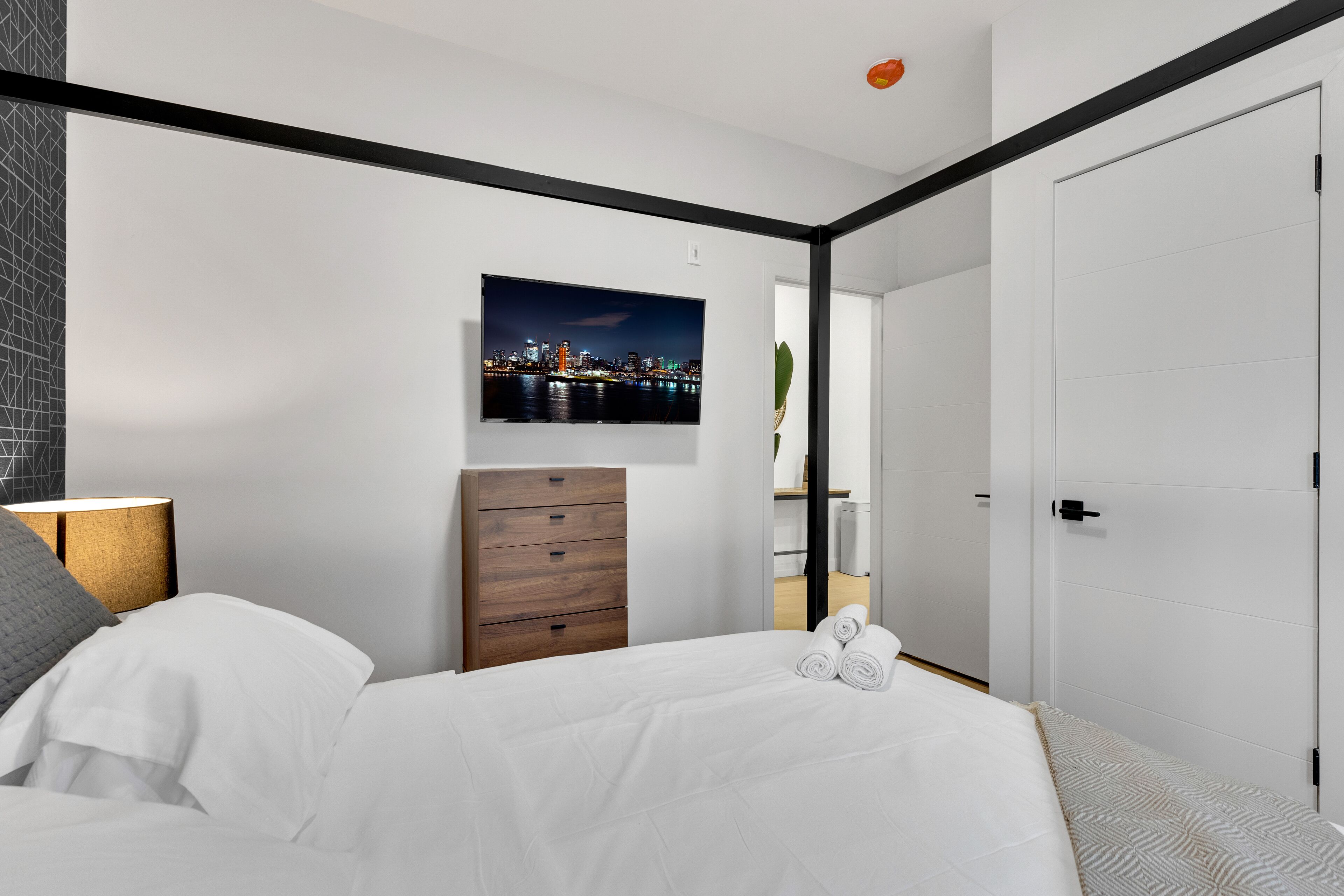 A modern bedroom with a bed, a nightstand, a wooden dresser, a wall-mounted TV, and a door.