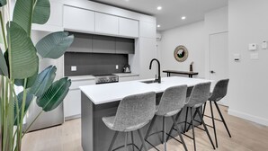 Een moderne keuken met witte kasten, een centraal eiland met barkrukken, en een grote groene plant.