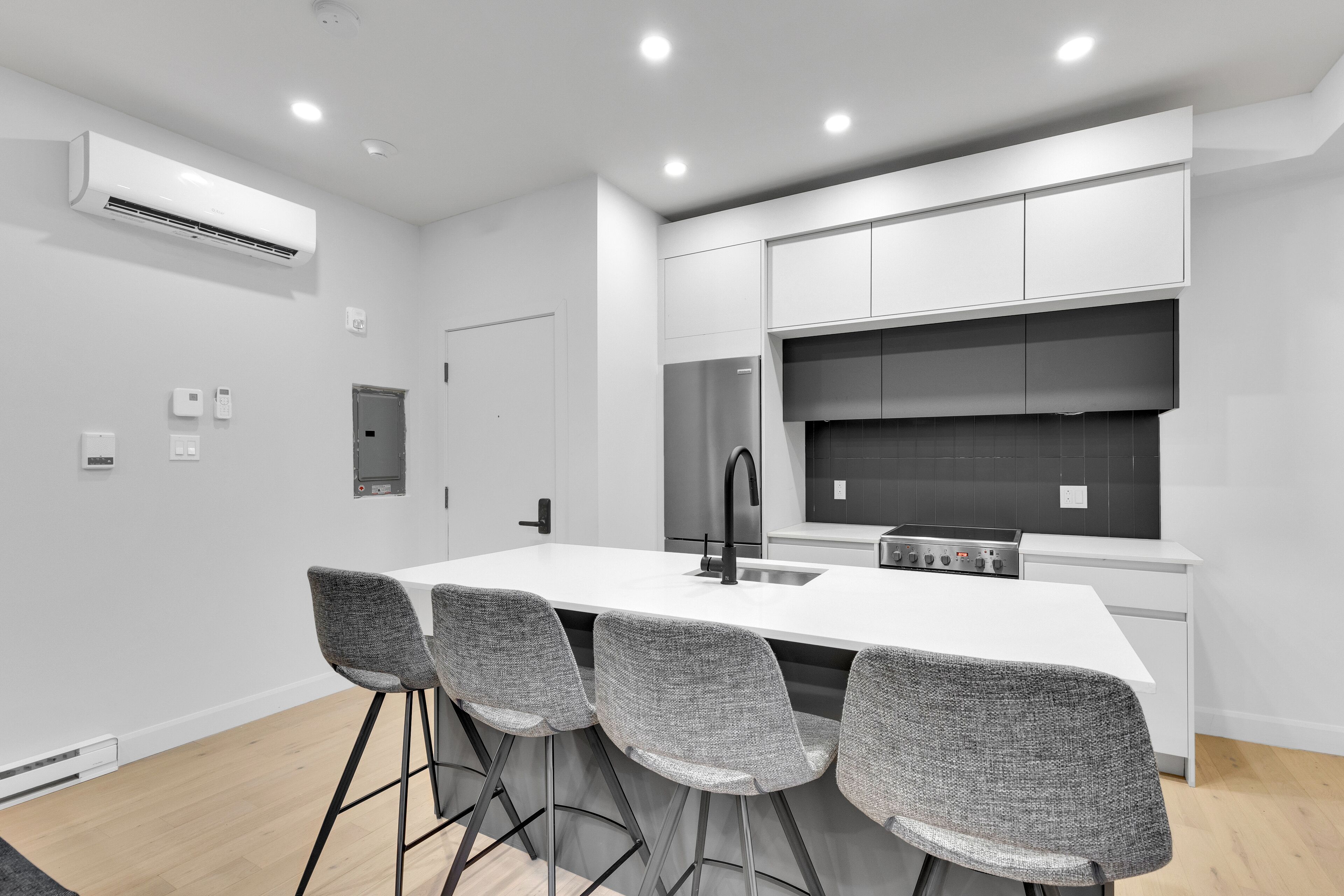 A modern kitchen with white cabinetry, a central island with bar stools, a stainless steel refrigerator, and a black backsplash.