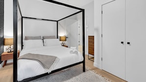 Een moderne slaapkamer met een hemelbed, een houten nachtkastje, een bijzettafeltje met een lamp en een witte kledingkast.