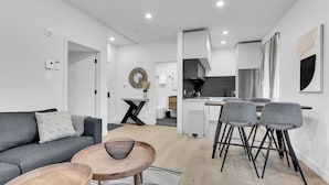 Een moderne woonkamer met een grijze bank, een houten salontafel en grijze fauteuils. In de achtergrond is een keuken met een eethoek zichtbaar.
