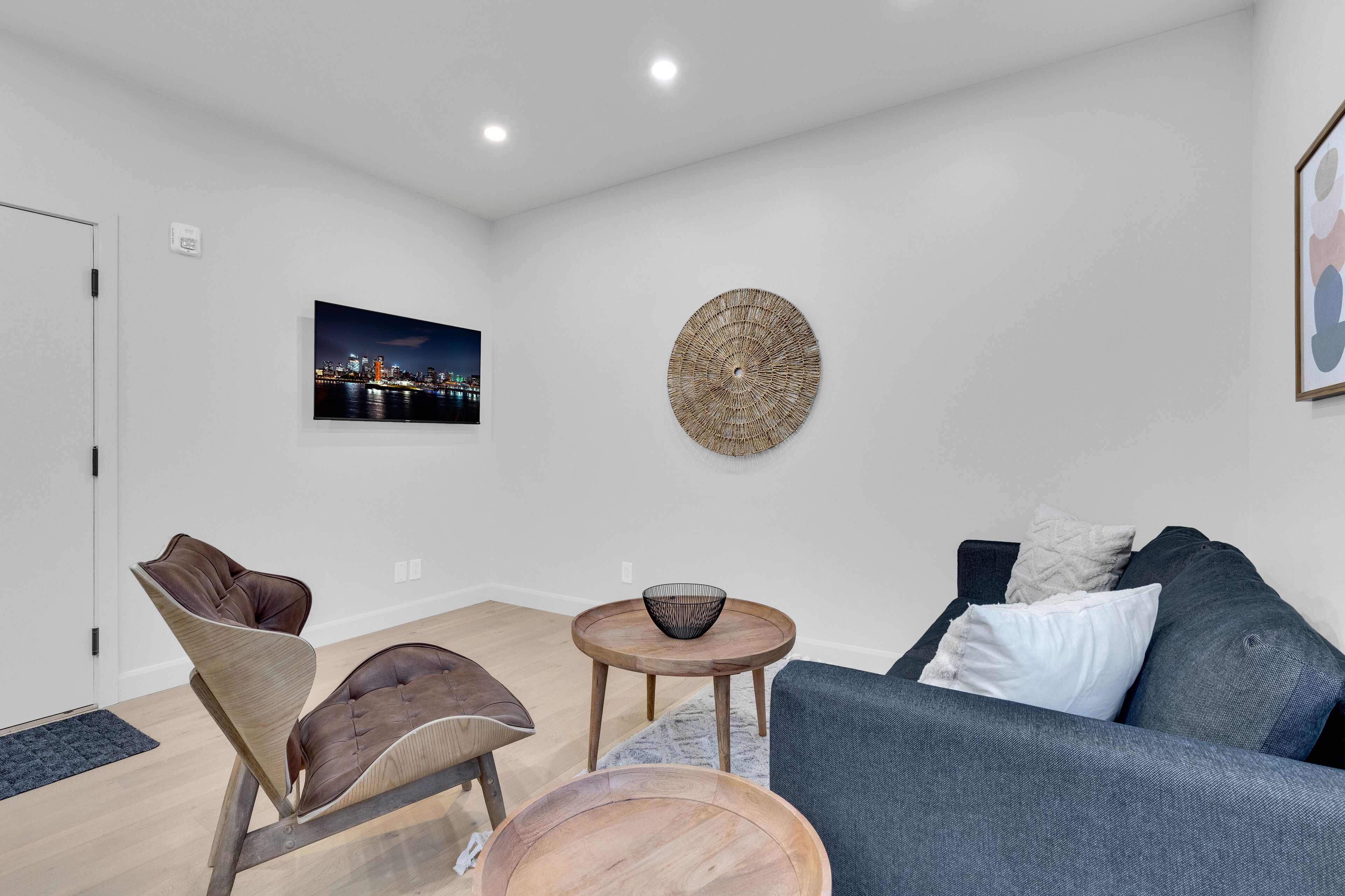 A modern living room with a grey sofa, a brown leather chair, a wooden coffee table, and a small round table with a wire basket.