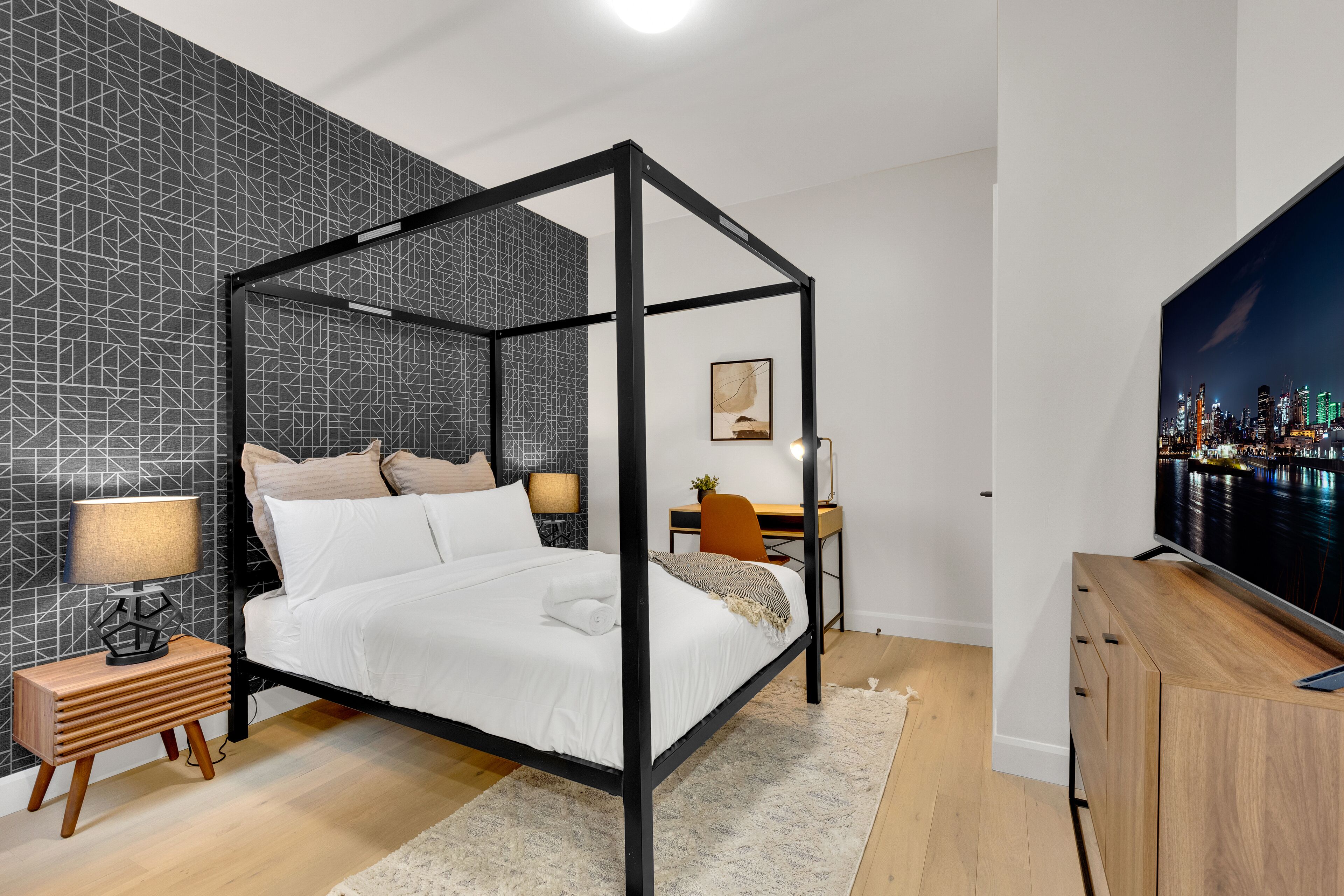 A modern bedroom with a four-poster bed, a cityscape TV, and a desk with a chair.