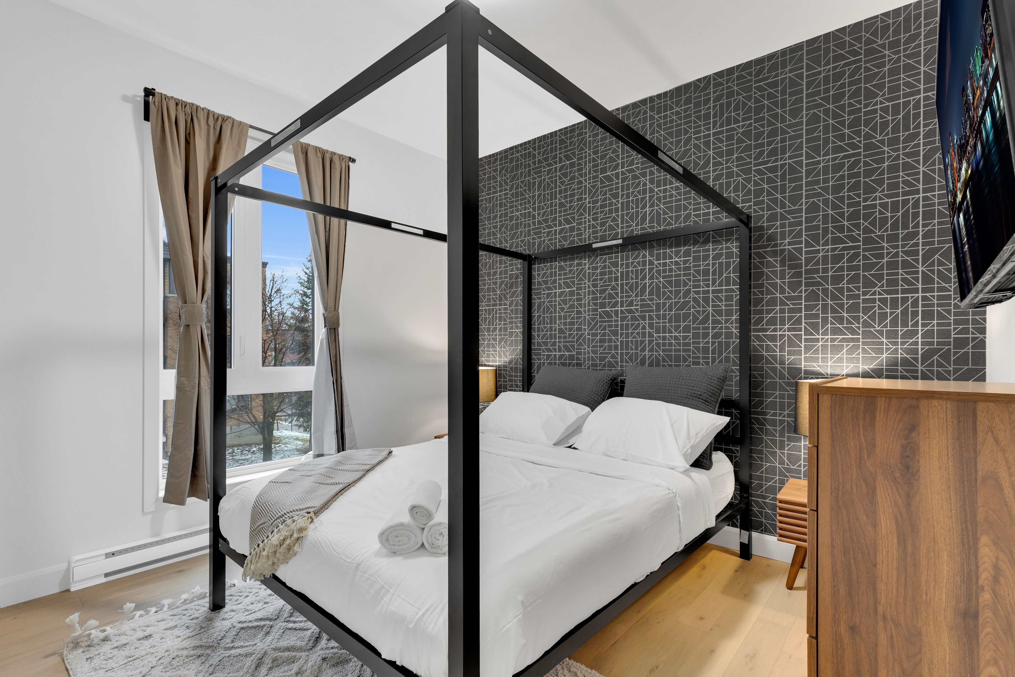 A modern bedroom with a four-poster bed, a geometric-patterned wall, and a wooden dresser.