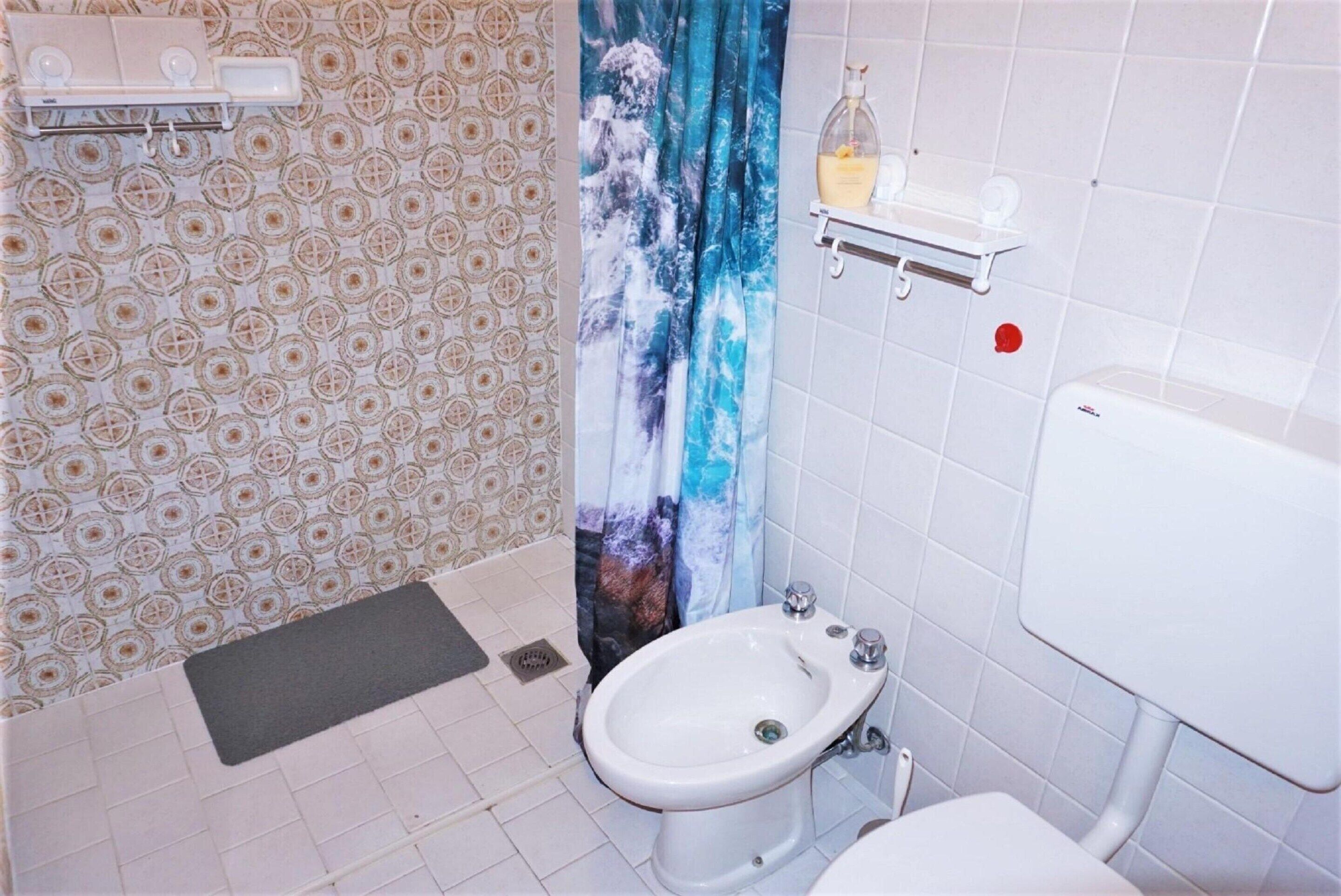 Une salle de bain équipée d’un bidet, de toilettes, d’un rideau de douche et d’une étagère contenant des articles de toilette.