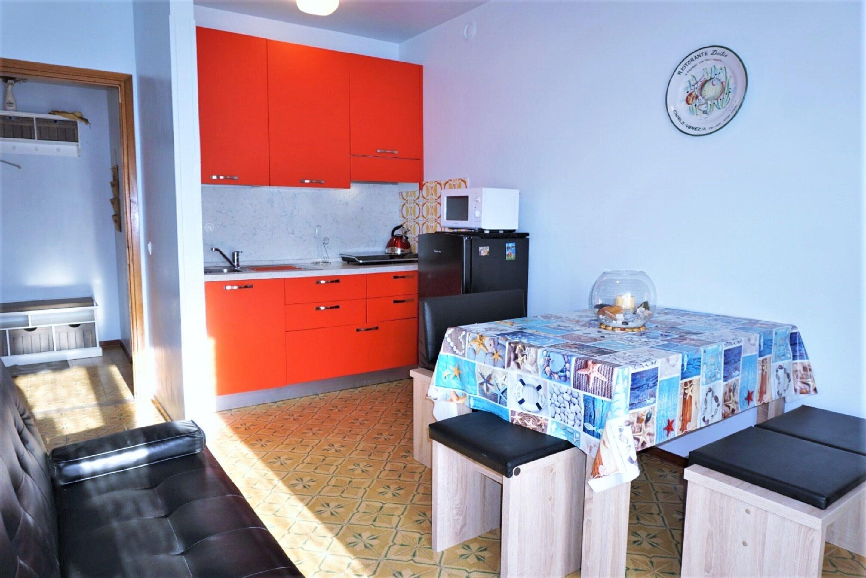 A compact kitchen with red cabinets, a microwave, and a refrigerator. There is a dining table with a colorful tablecloth and benches.