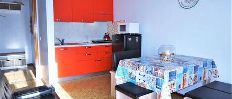 A compact kitchen with red cabinets, a microwave, and a refrigerator. There is a dining table with a colorful tablecloth and benches.