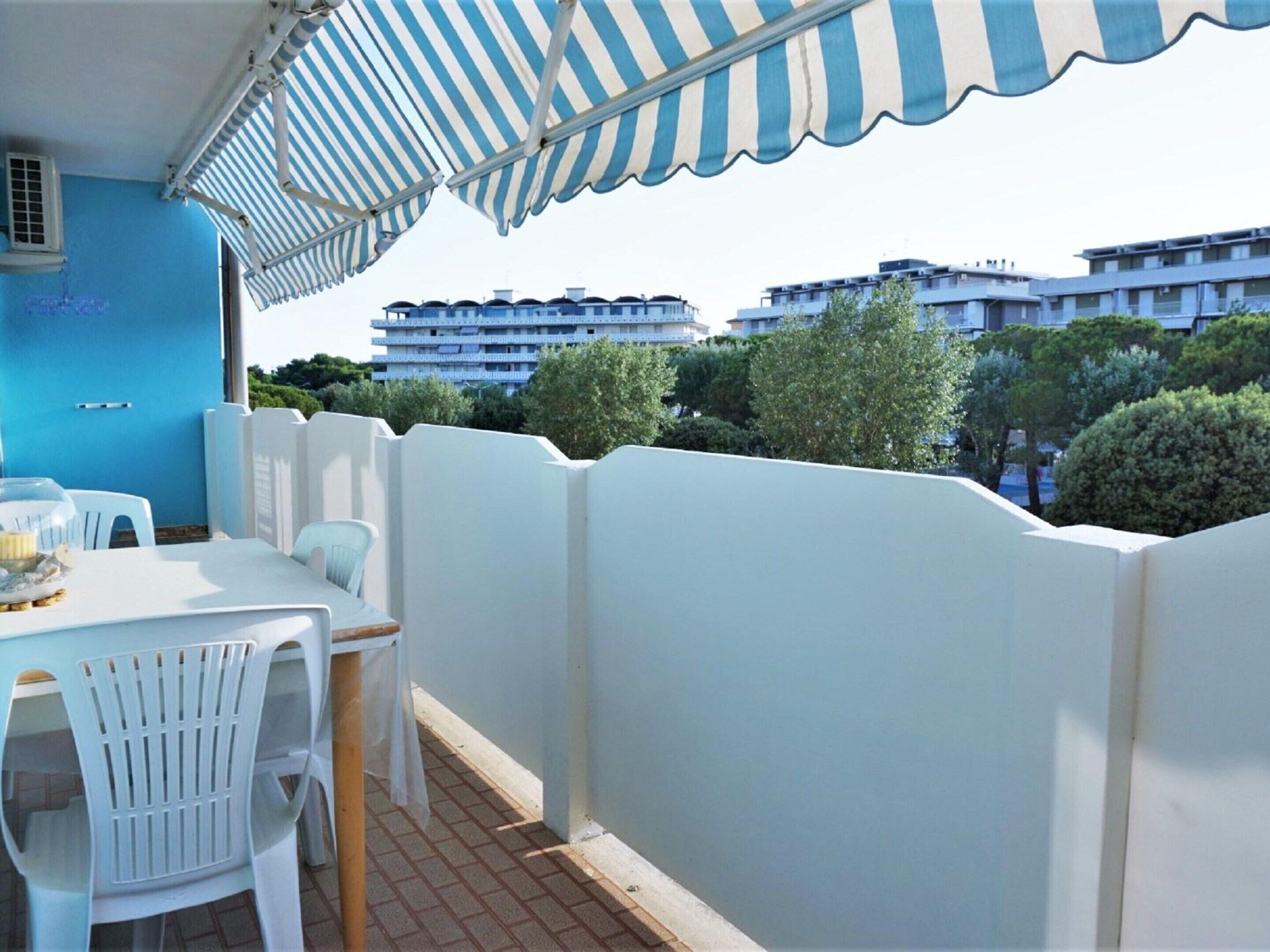 A balcony with a striped awning, white plastic chairs, and a table with a glass container.