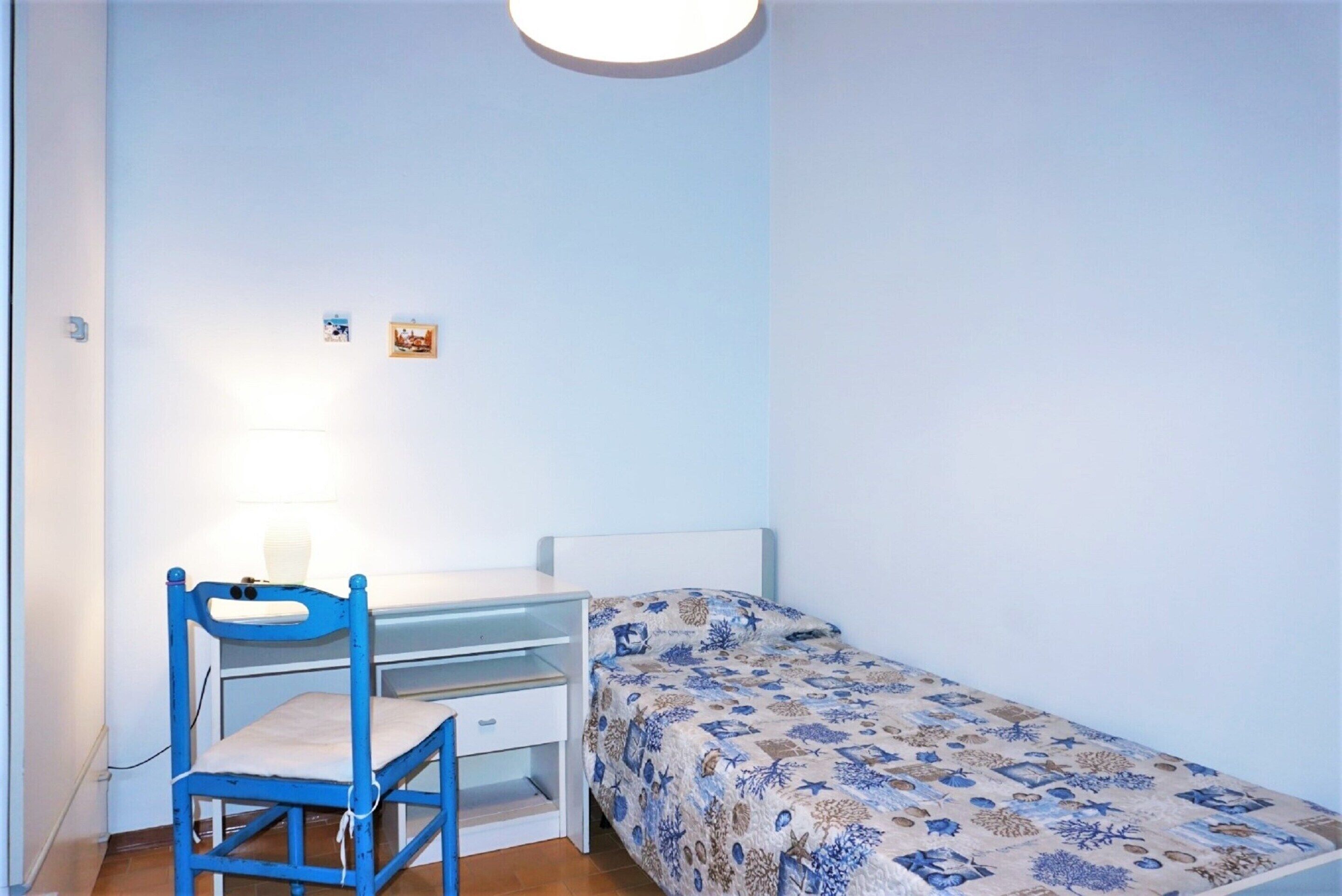 Een kleine kamer met een enkel bed, een bureau, een stoel, een lamp en twee kleine decoraties aan de muur.