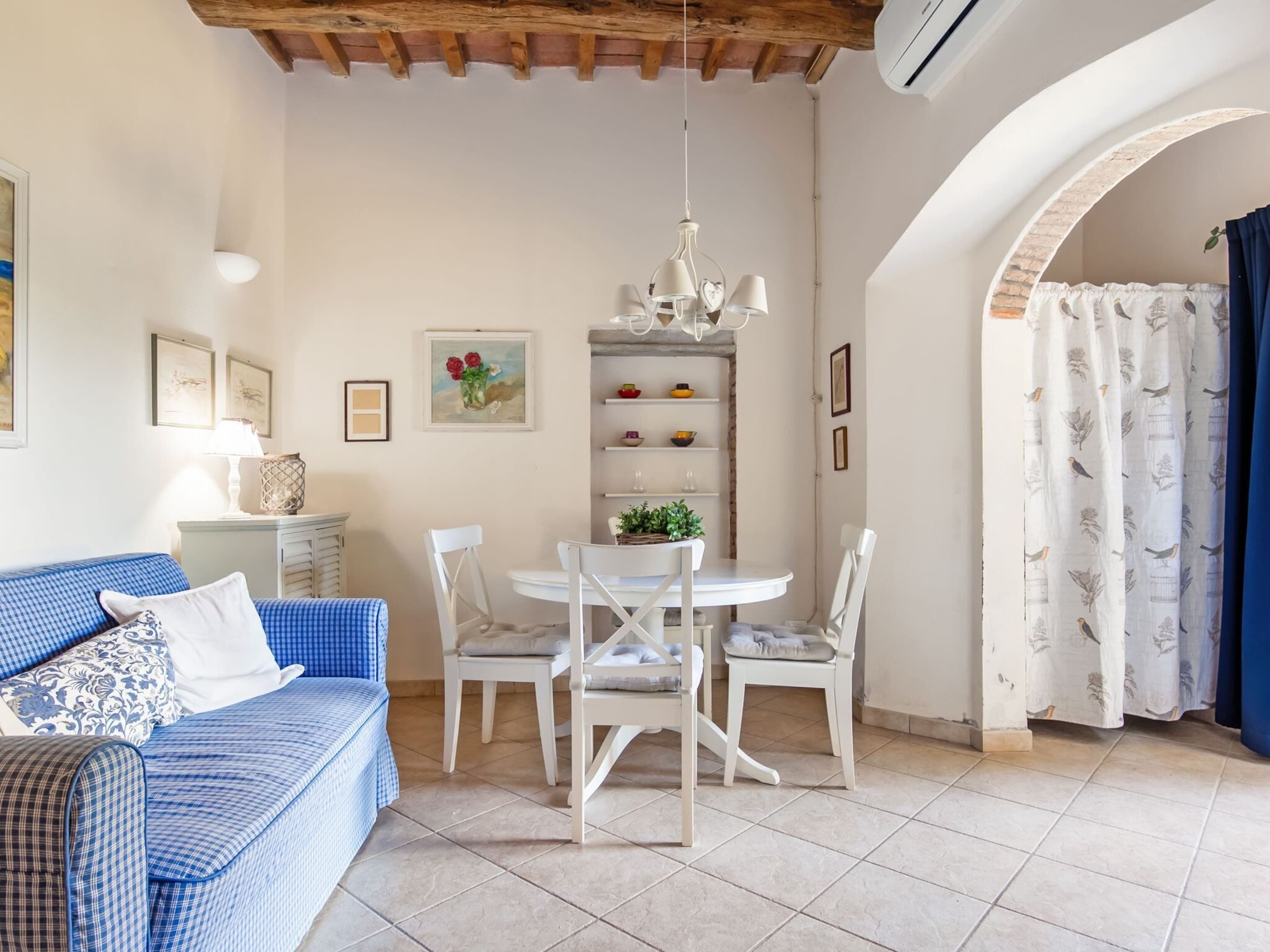 Un salon chaleureux avec un canapé bleu à carreaux, des chaises blanches, une table à manger surmontée d’un lustre et un coin cuisine avec des étagères.