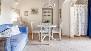 Ein gemütliches Wohnzimmer mit einem blau karierten Sofa, weißen Stühlen, einem Esstisch mit einer Pendelleuchte und einem Küchenbereich mit Regalen.